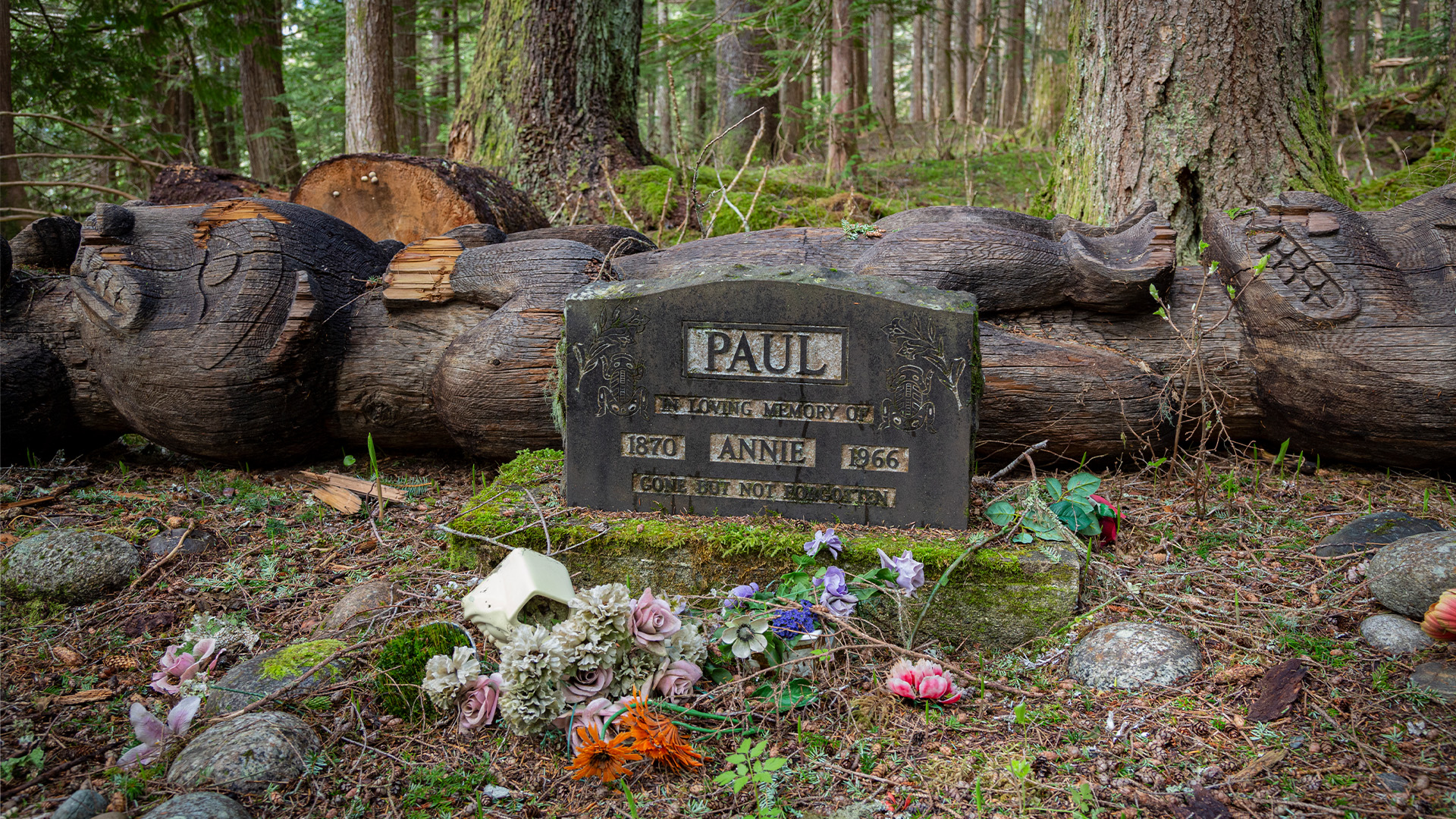 En gravsten framför en liggande totempåle. Fotografi.